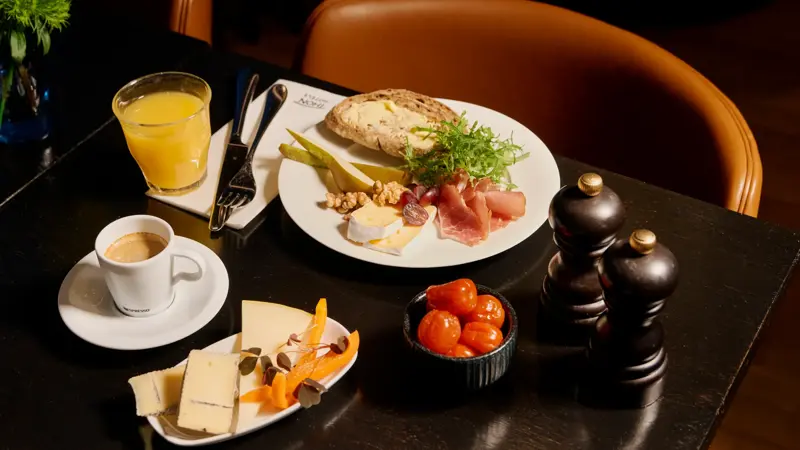 En frokosttallerken med grovt brød med smør, spekeskinke, ost, pære, valnøtter og salat. Ved siden av er et glass appelsinjuice, en kopp espresso og en tallerken med ulike oster.