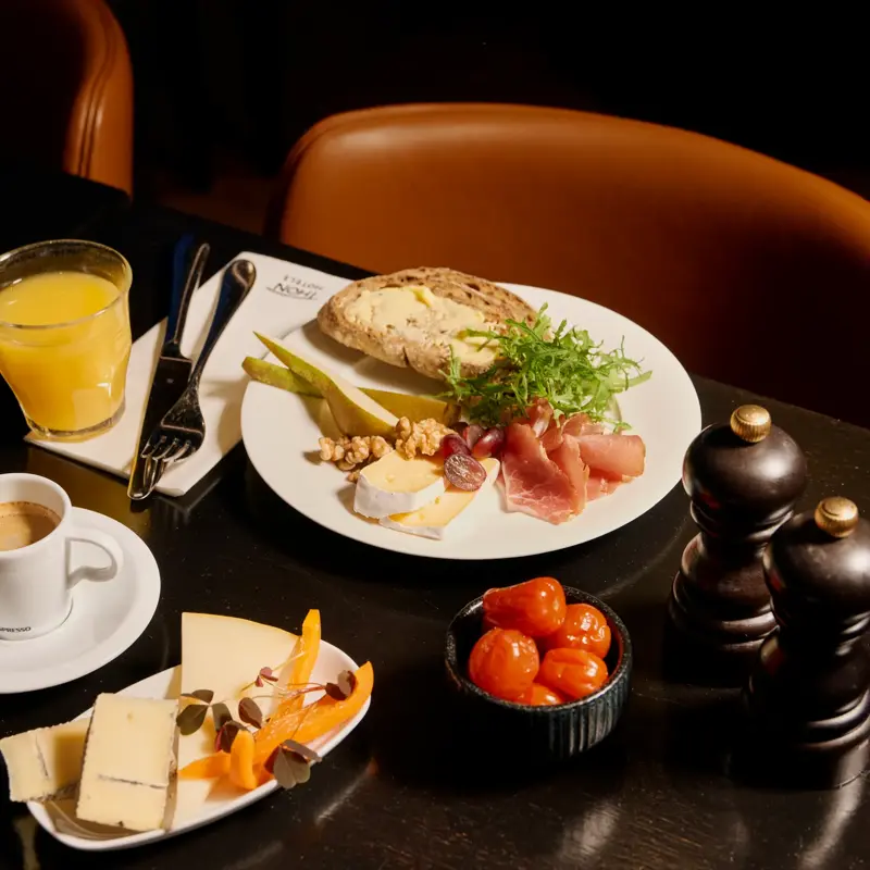 En frokosttallerken med grovt brød med smør, spekeskinke, ost, pære, valnøtter og salat. Ved siden av er et glass appelsinjuice, en kopp espresso og en tallerken med ulike oster.