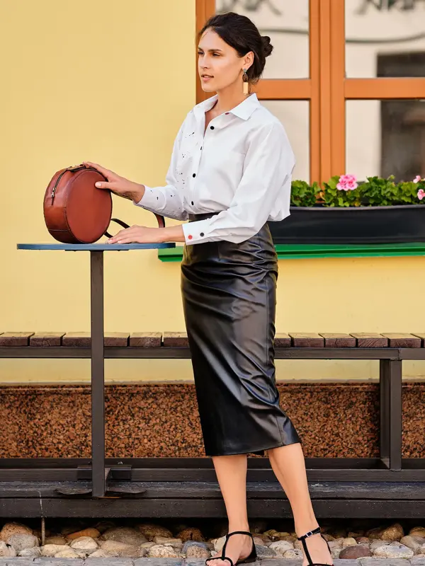 Dame med sort skinnskjørt, hvit skjorte, sorte sandaler og brun veske står ved et bord utendørs 