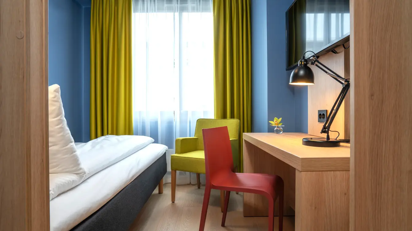 Enkelt seng, blå vegger, gul stol og gule gardiner, skrivebord og TV på enkeltrom på Thon Hotel Astoria