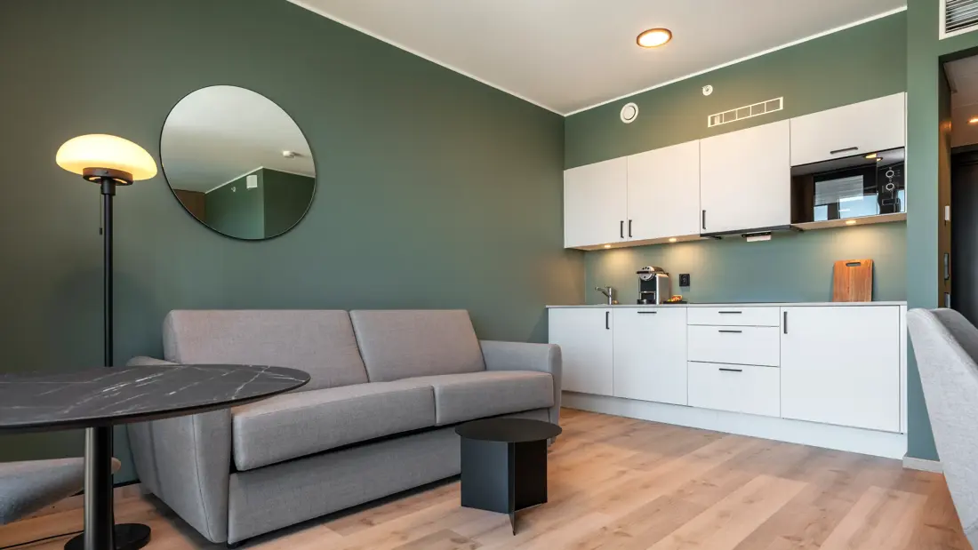 Superior rom med kjøkken krok, sofa, grønne vegger, parkett, og rundt speil på veggen 