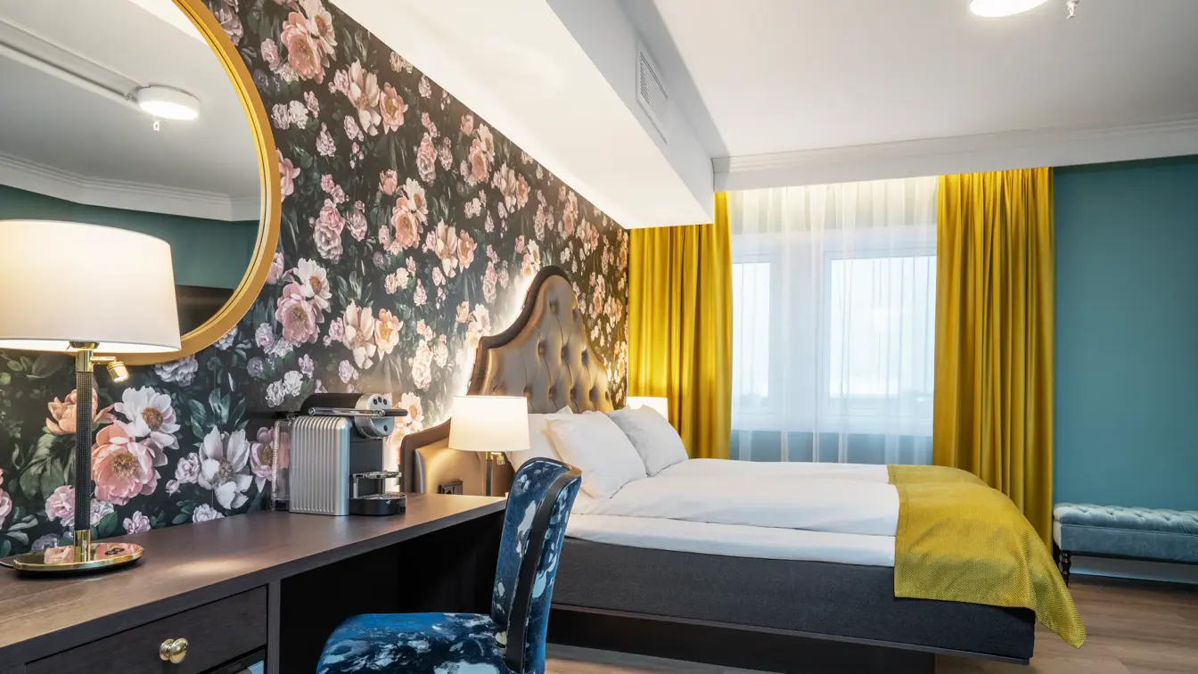 Mørk tapet med store lyserosa roser. Vindu med gule gardiner fra tak til gulv. Dobbeltseng med gult sengeteppe. Mørkt skrivebord med kaffemaskin, speil over og mønstrete stol. I superior rom på Thon Hotel Høyers i Skien.