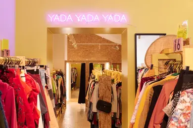 Interiør fra en bruktbutikk med fargerike klær og et neoenskilt på veggen som sier Yada Yada Yada