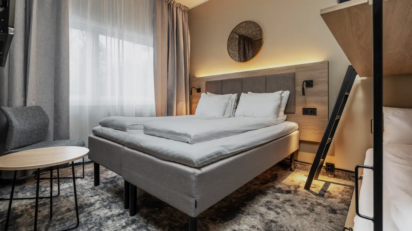 Dobbeltrom med queen seng, lys rundt sengegavl, lite bord med stol, stort vindu og lange gardiner