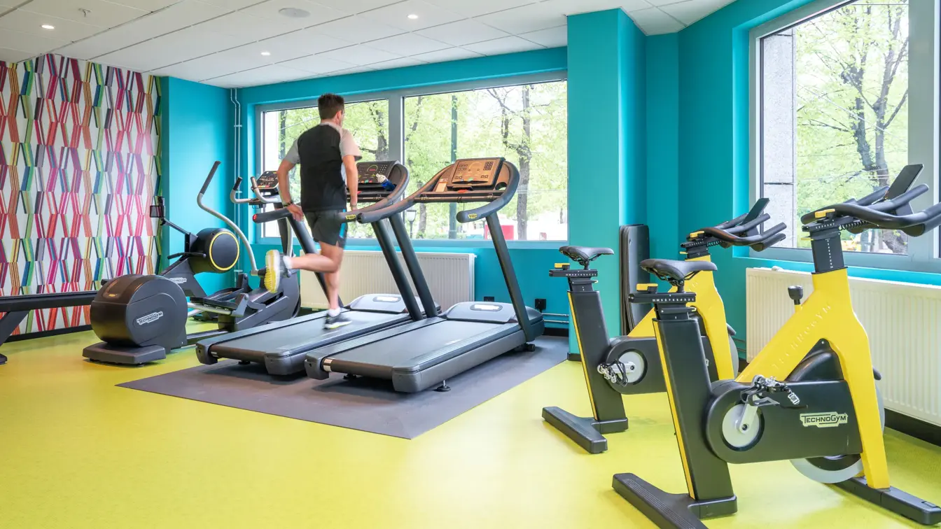 Mann som løper på tredemølle i treningsrom med gult gulv, turkise vegger, fargerik tapet og store vinduer med utsikt på Thon Hotel Cecil.