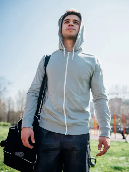 Mann med grå hettejakke, sort bukse og sort treningsbag på skulderen poserer for kamera 