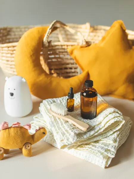 Moses basket, hair brush, babies goods diaper, moon and linen star shaped cushions, dinosaur toy, cream, shampoo or oil on beige background with copy space. Newborn unisex baby necessities and clothing on cozy nursery in morning sunlights. Baby shower concept.