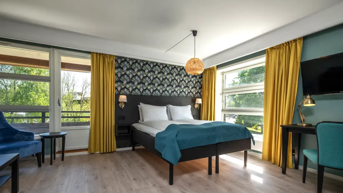 Superior rom med dobbeltseng, mønstrete tapet bak sengen, store vinduer og gule gardiner, vegghengt TV og skrivebord
