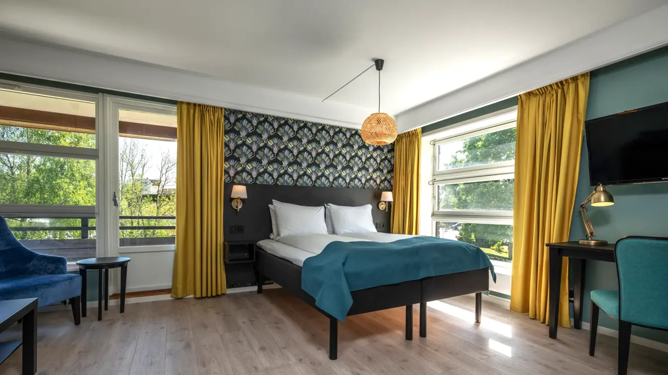 Superior rom med dobbeltseng, mønstrete tapet bak sengen, store vinduer og gule gardiner, vegghengt TV og skrivebord
