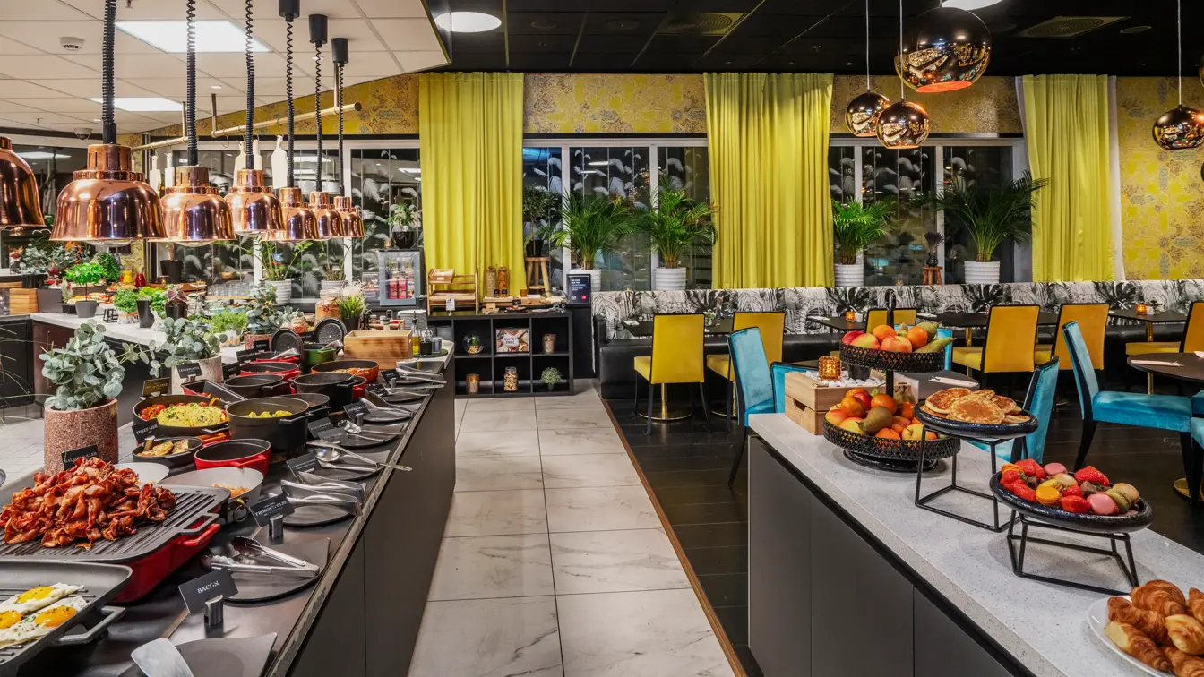 Frokostbuffet med diverse varmmat på venstre side og frukt på høyre side. Gule gardiner, gul tapet, småbord med blå og gule stoler.