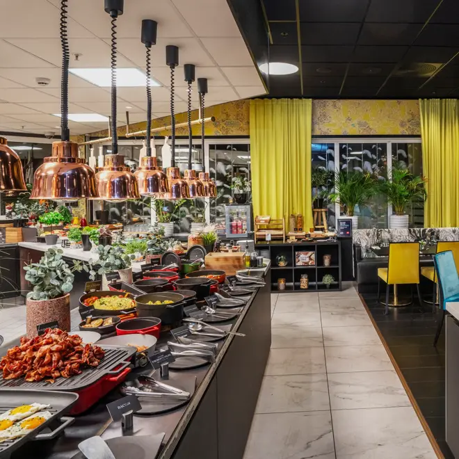 Frokostbuffet med diverse varmmat på venstre side og frukt på høyre side. Gule gardiner, gul tapet, småbord med blå og gule stoler.