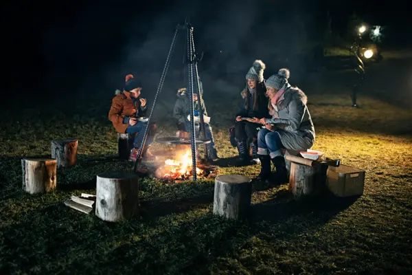 Mother and three kids sitting around a bonfire in the backyard. There is a grill over the bonfire where some sausages are roasting over fire. Family is eating the grilled sausages and talking.
Shot with Canon R5