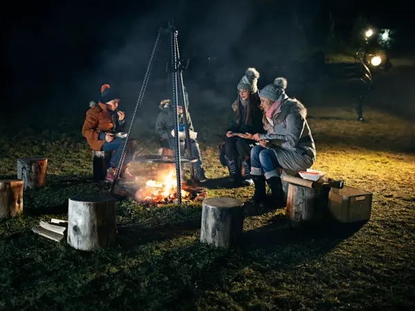 Mother and three kids sitting around a bonfire in the backyard. There is a grill over the bonfire where some sausages are roasting over fire. Family is eating the grilled sausages and talking.
Shot with Canon R5