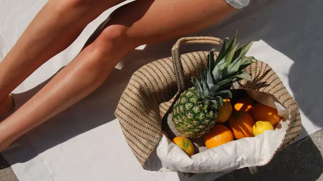 Nærbilde av solbrune legger og strandveske med frukt 