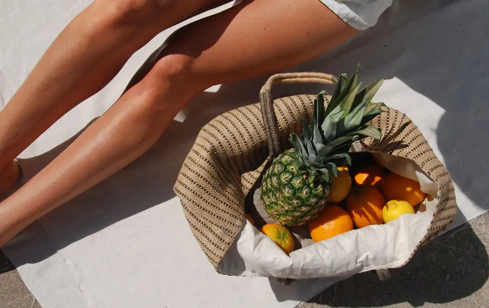 Nærbilde av solbrune legger og strandveske med frukt 