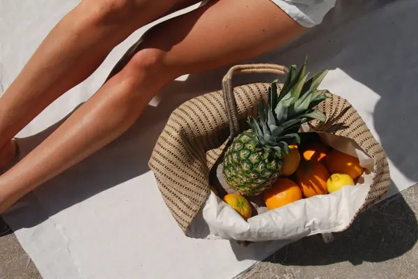 Nærbilde av solbrune legger og strandveske med frukt 