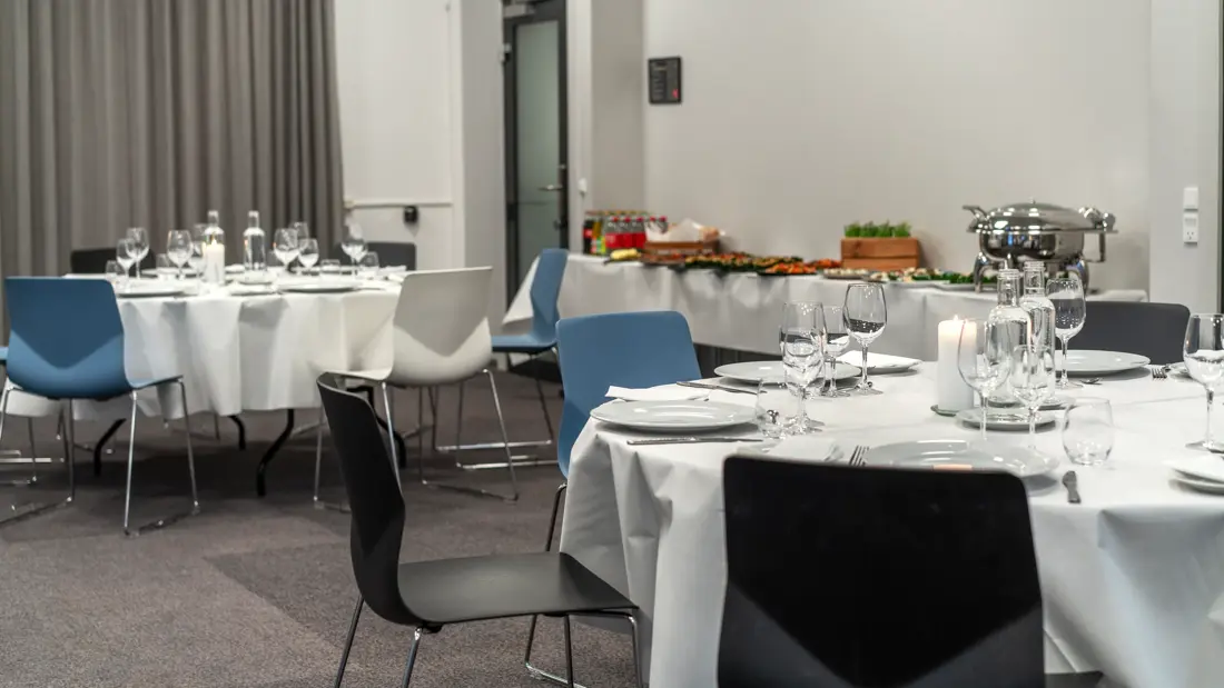 Selskapslokale med bankettoppsett. Runde spisebord dekket med hvit duk, hvite tallerkener, bestikk, glass, vannkarafler, kubbelys. Seks stoler rundt hvert bord, ensfarget i sort, hvit eller blått. I bakgrunnen ses langbord med drikke, tapasretter.