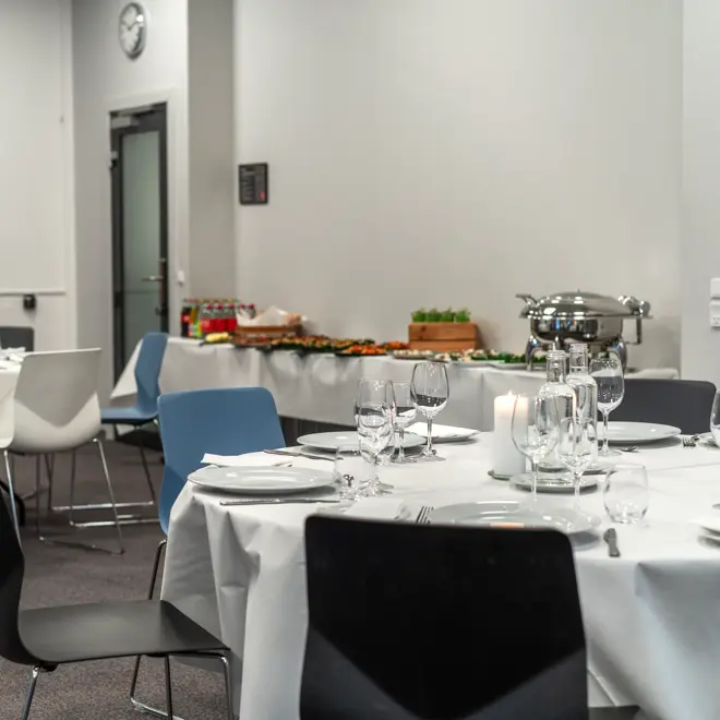 Selskapslokale med bankettoppsett. Runde spisebord dekket med hvit duk, hvite tallerkener, bestikk, glass, vannkarafler, kubbelys. Seks stoler rundt hvert bord, ensfarget i sort, hvit eller blått. I bakgrunnen ses langbord med drikke, tapasretter.