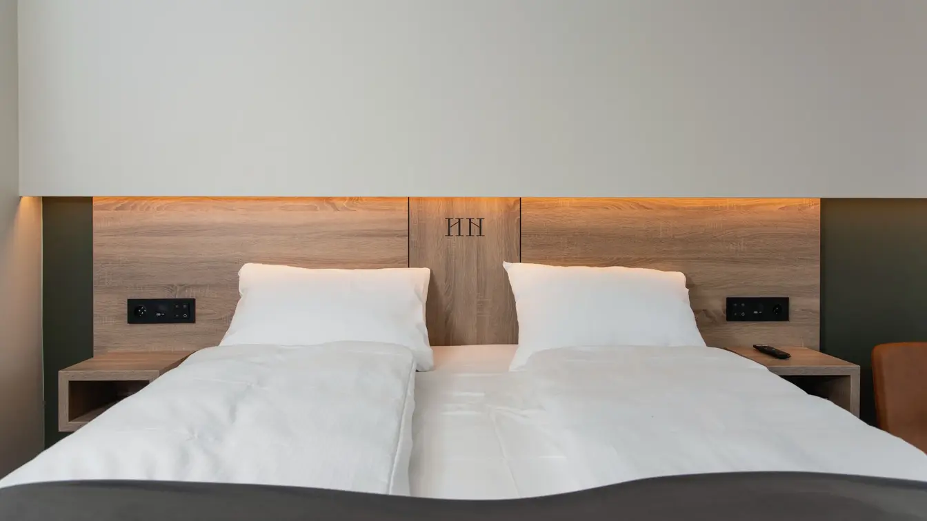 Dobbeltseng med sengegavl i lyst træ og hvit vegg. Hvite laken og mørkt sengeteppe. På Thon Hotel Norge i Kristiansand.
