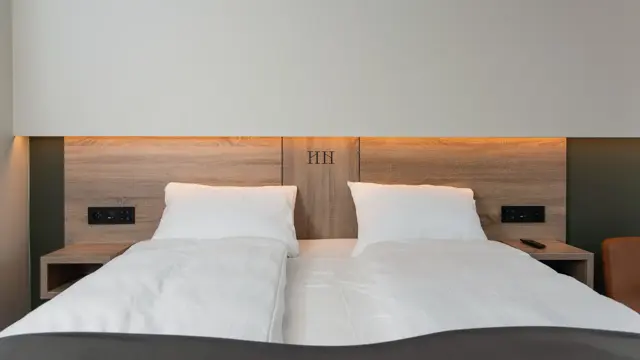 Dobbeltseng med sengegavl i lyst træ og hvit vegg. Hvite laken og mørkt sengeteppe. På Thon Hotel Norge i Kristiansand.