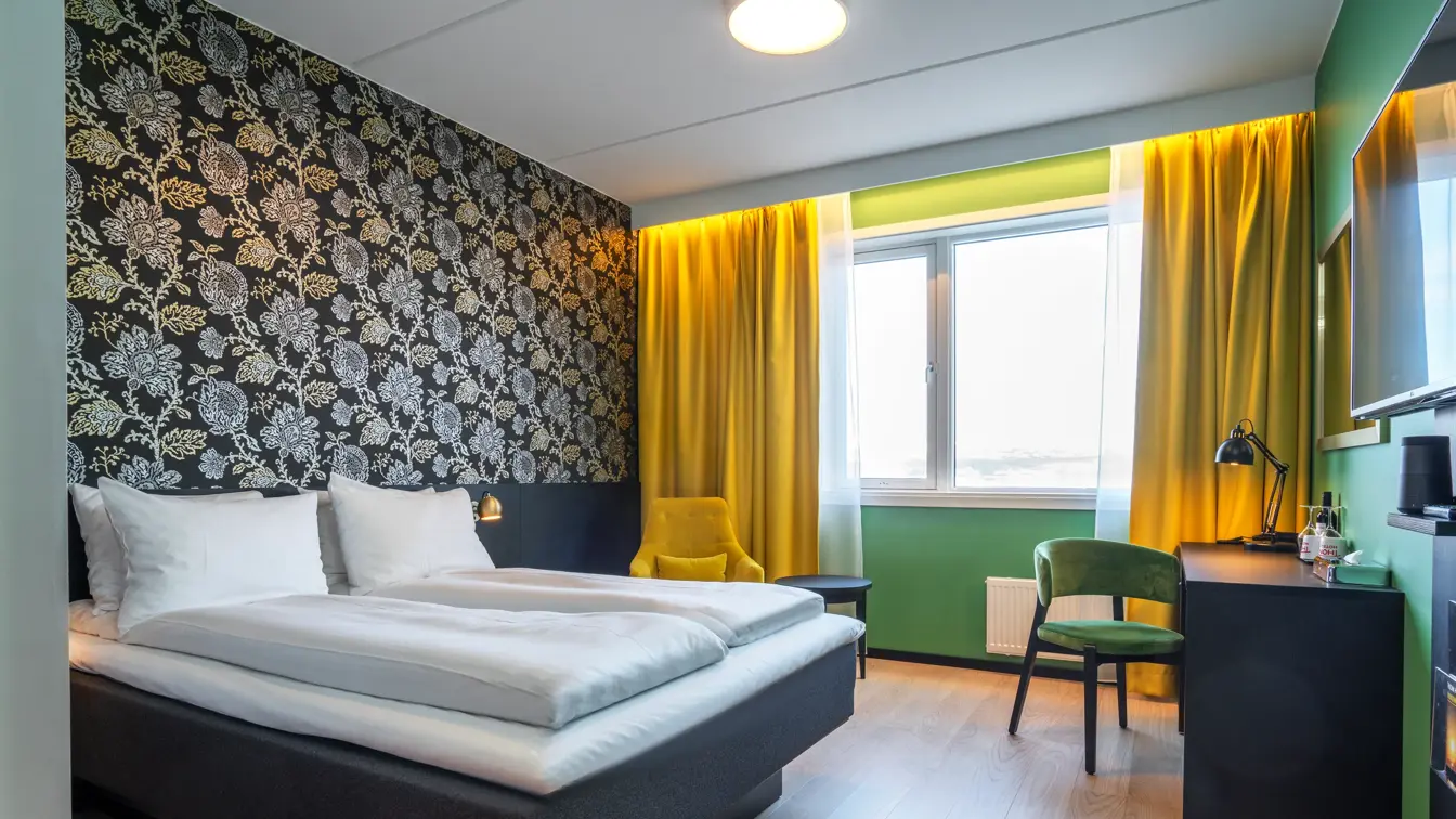 Dobbeltseng, mørk mønstrete tapet med blomster, gule gardiner, grønne vegger, skrivebord grønn stol og TV på dobbeltrom på Thon Hotel Kirkenes