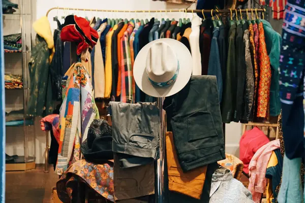 Klær på bord og stativ i butikk. Cowboyhatt, bukser og fargerike jakker.