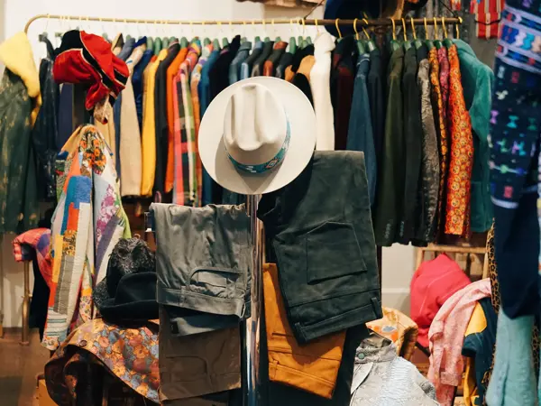 Klær på bord og stativ i butikk. Cowboyhatt, bukser og fargerike jakker.