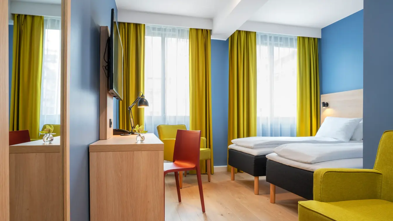 Gul stol, separate senger, blå vegger, gule gardiner og skrivebord på standard rom twin på Thon Hotel Astoria