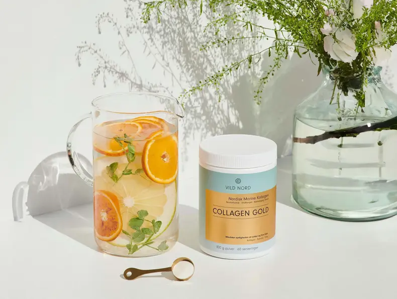 En boks med Collagen Gold fra Vild Nord står ved siden av et glass med vann og skiver av appelsin og sitron.