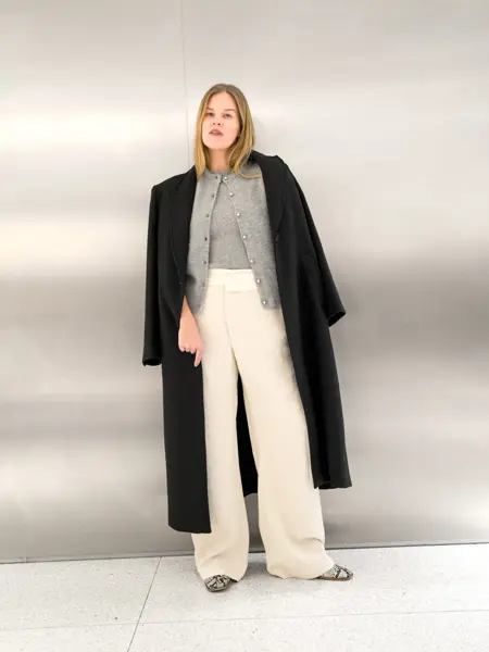 Dame i mørk kåpe, grå cardigan og topp og hvit bukse