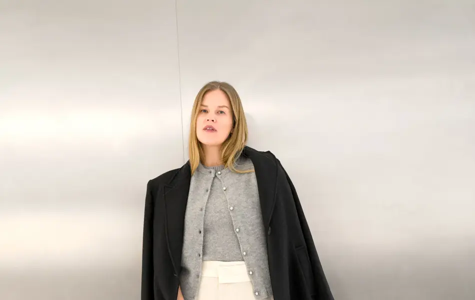 Dame i mørk kåpe, grå cardigan og topp og hvit bukse