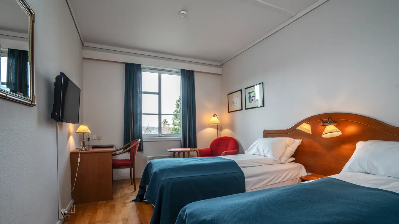 To enkelsenger med blå sengetepper, skrivebord med vegghengt TV i bakgrunnen