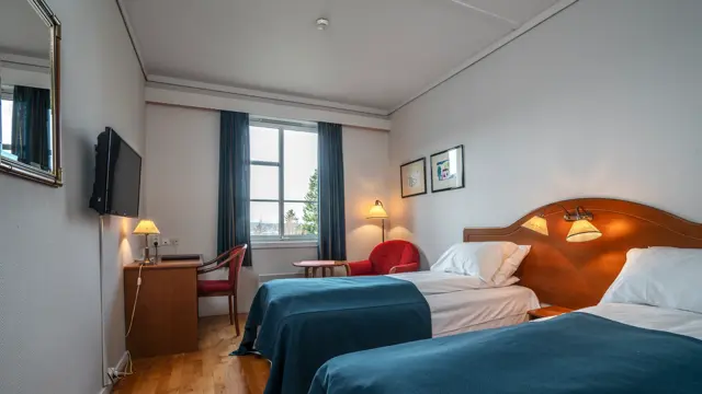 To enkelsenger med blå sengetepper, skrivebord med vegghengt TV i bakgrunnen