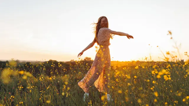 Dame med lang blomstrete kjole danser i gul blomstereng i vårsol 