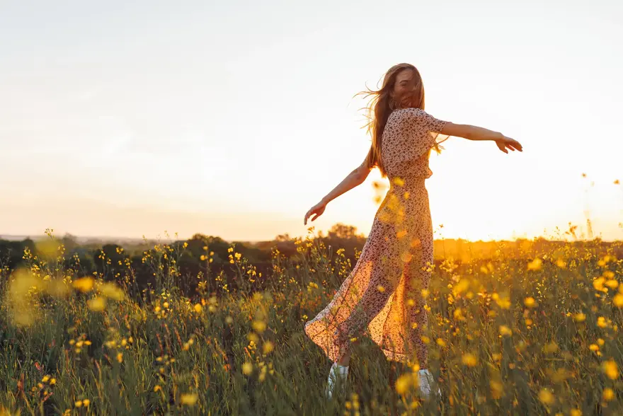 Dame med lang blomstrete kjole danser i gul blomstereng i vårsol 