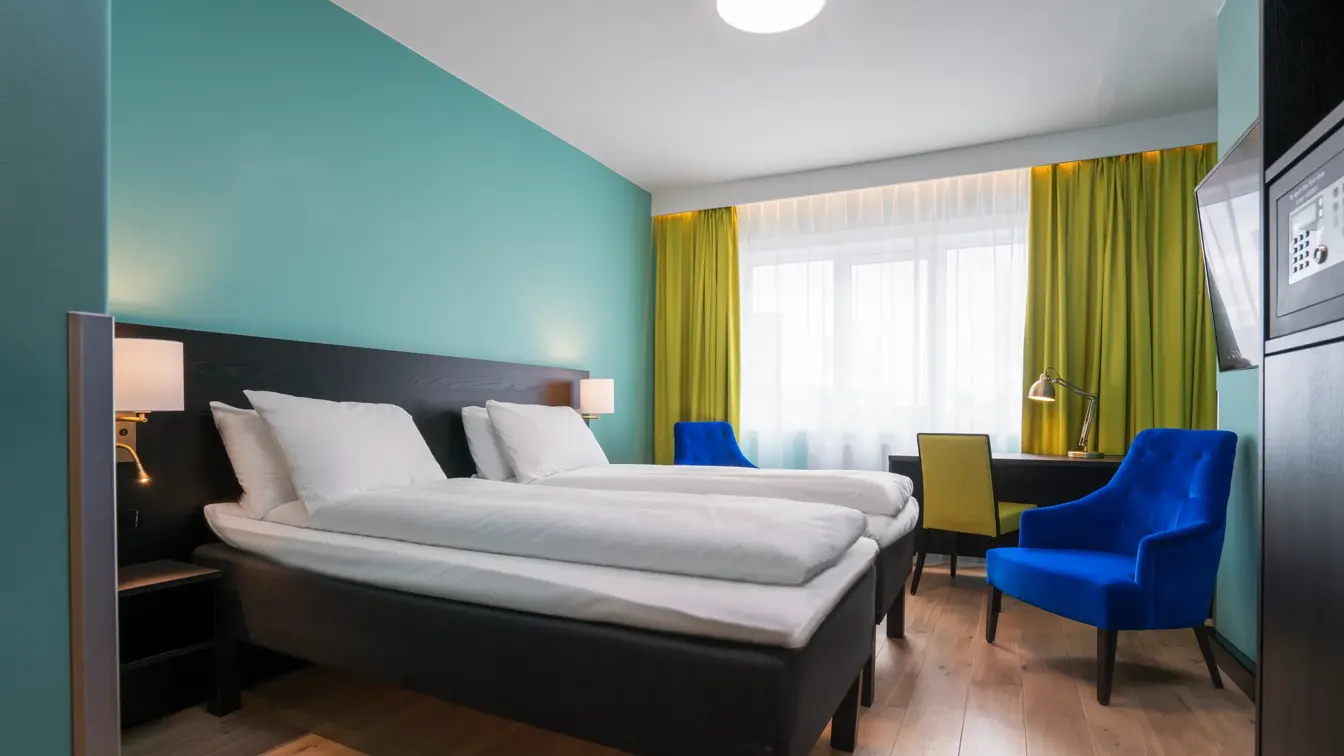 Twin room med hvit sengetøy, gule gardiner, skrivepult og blå lenestol på Thon Hotel Europa i Oslo sentrum rett ved Slottsparken