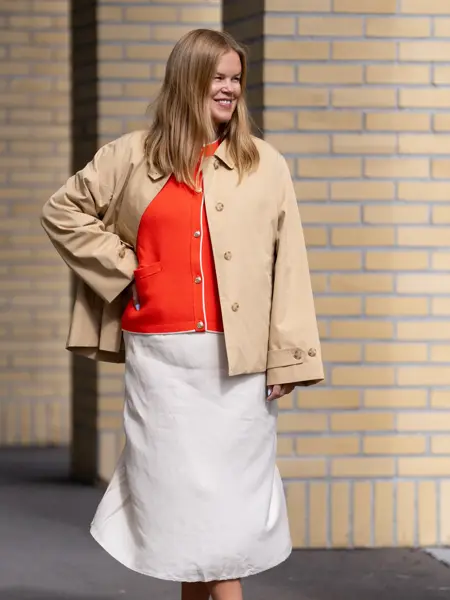 Dame med beige knelangt skjørt, rød topp og beige kort trenchjakke.