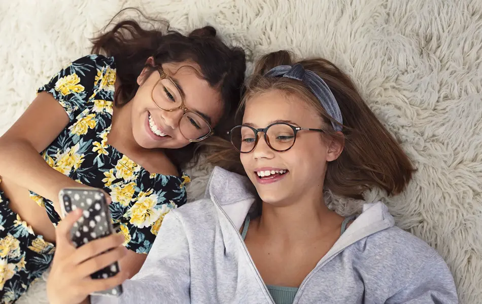 to unge jenter med briller som ligger på gulvet med mobil i hånden