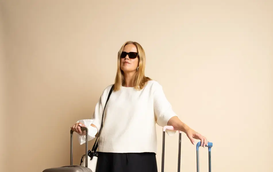 Dame modelle med sorte solbriller, hvit genser, sort skjørt og sorte sko, står med tre tillekofferter.