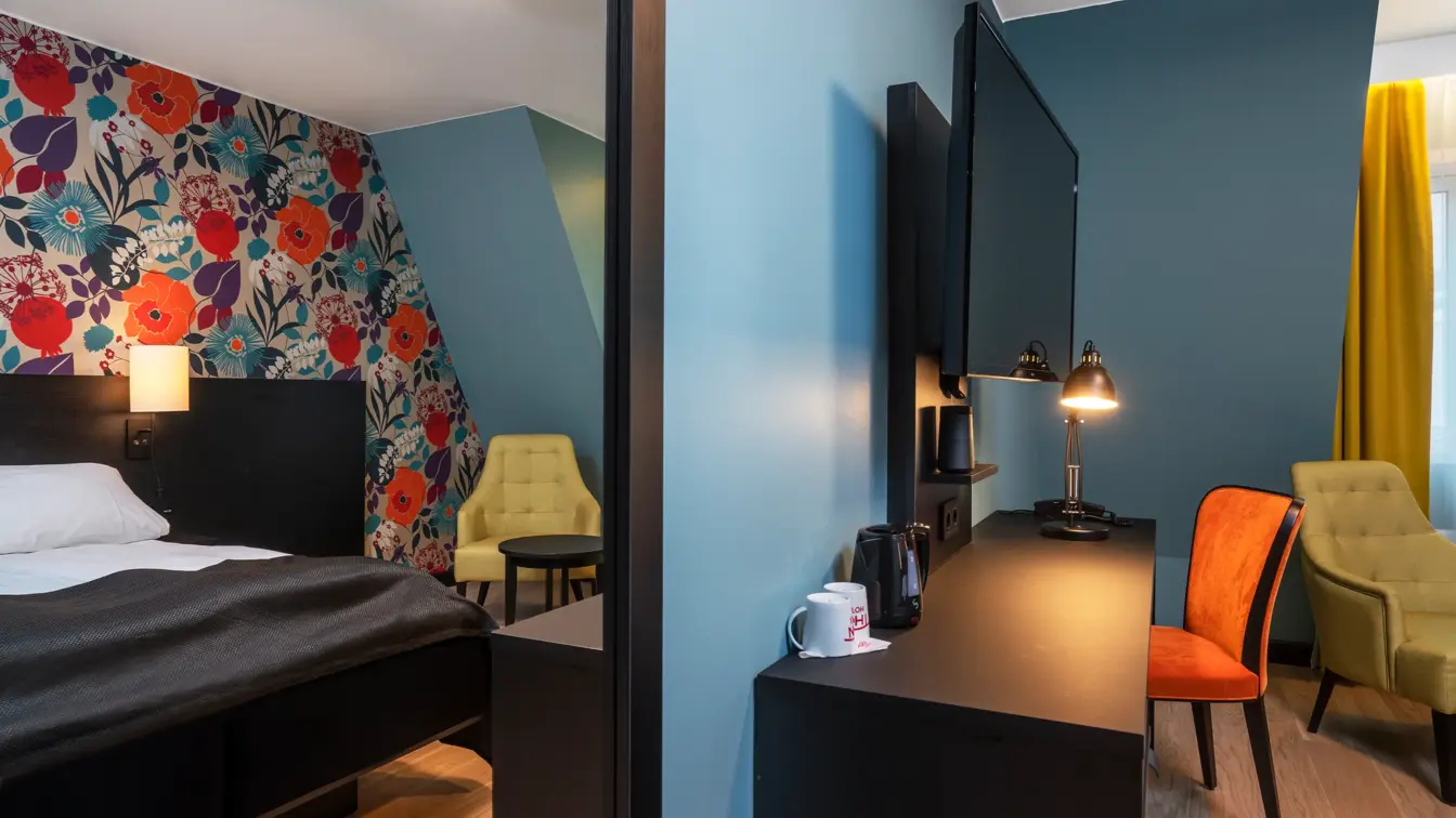 Blomstrete tapetvegg med blå vegger, gule gardiner, dobbeltseng, gul stol, svart skrivedbord, oransje stol og en flatskjerm hengende på vegg.