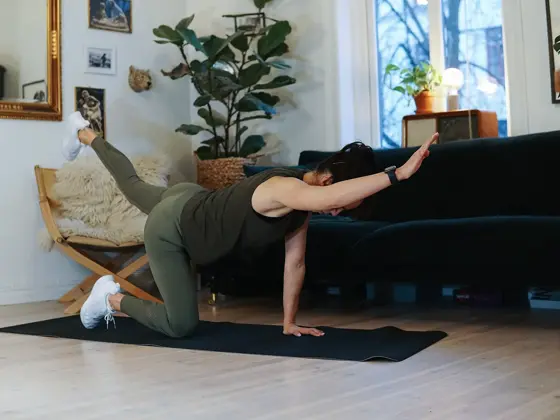 treningsekspert Anna Johansen trener på matte på gulvet, mørkegrønn sofa i bakgrunnen, lys stol med pels oppi, str grønn plante, bilder på veggen