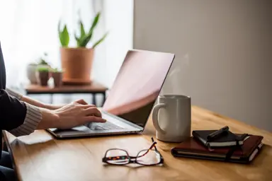 En laptop, et par briller, en kaffekopp og to notatbøker er på et skrivebord, med to hender som skriver på laptopen