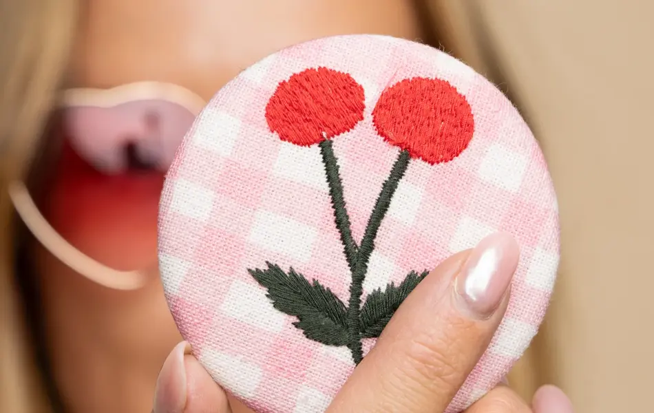 Nærbilde av hånd som holder et lite speil med rosa stoff og broderi på cover.