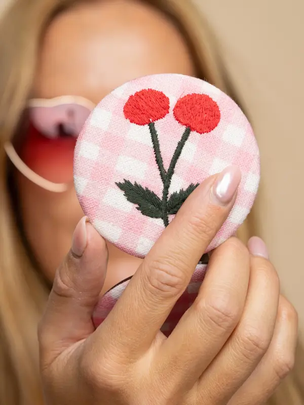 Nærbilde av hånd som holder et lite speil med rosa stoff og broderi på cover.