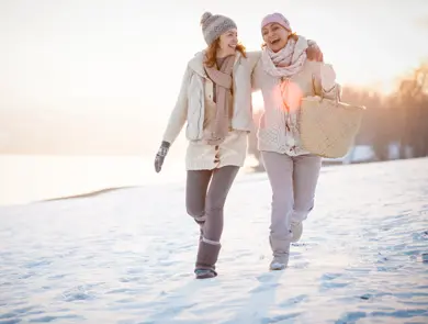Two happy women are embracing while walking on snow-covered field on a sunny day.