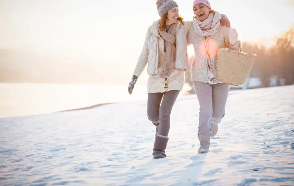 Two happy women are embracing while walking on snow-covered field on a sunny day.