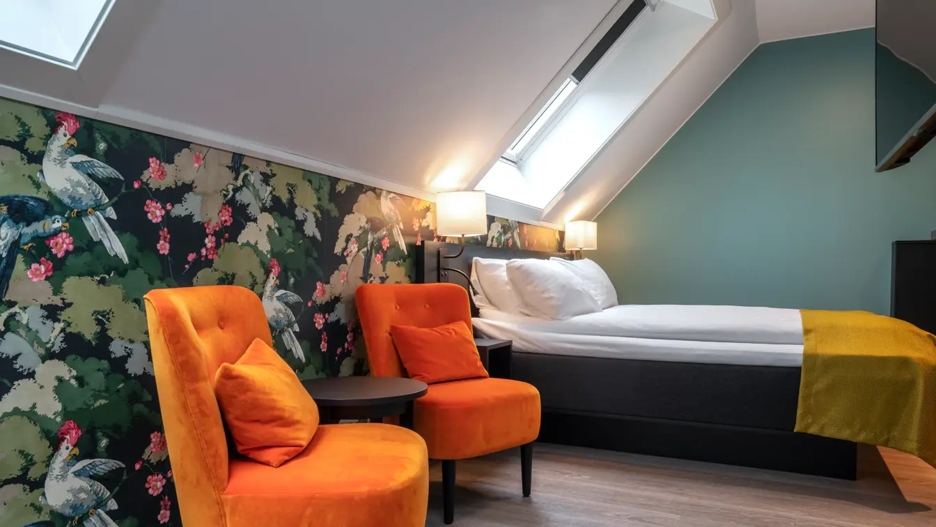 Tapet med blomster og fugler, to oransje stoler med tilhørende avlastningsbord, to takvinduer, parkettgulv, en dobbeltseng og TV.