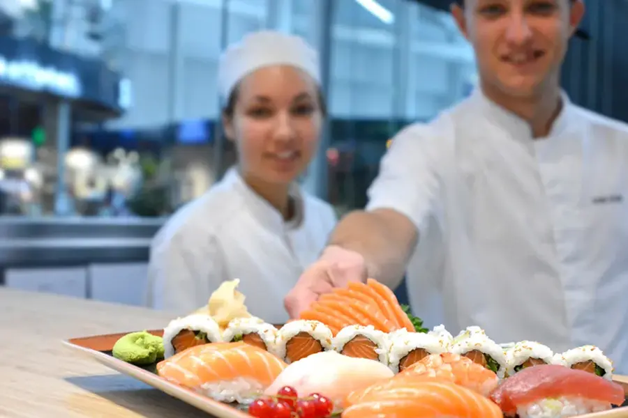 To sushi kokk som smiler og serverer sushi på benken