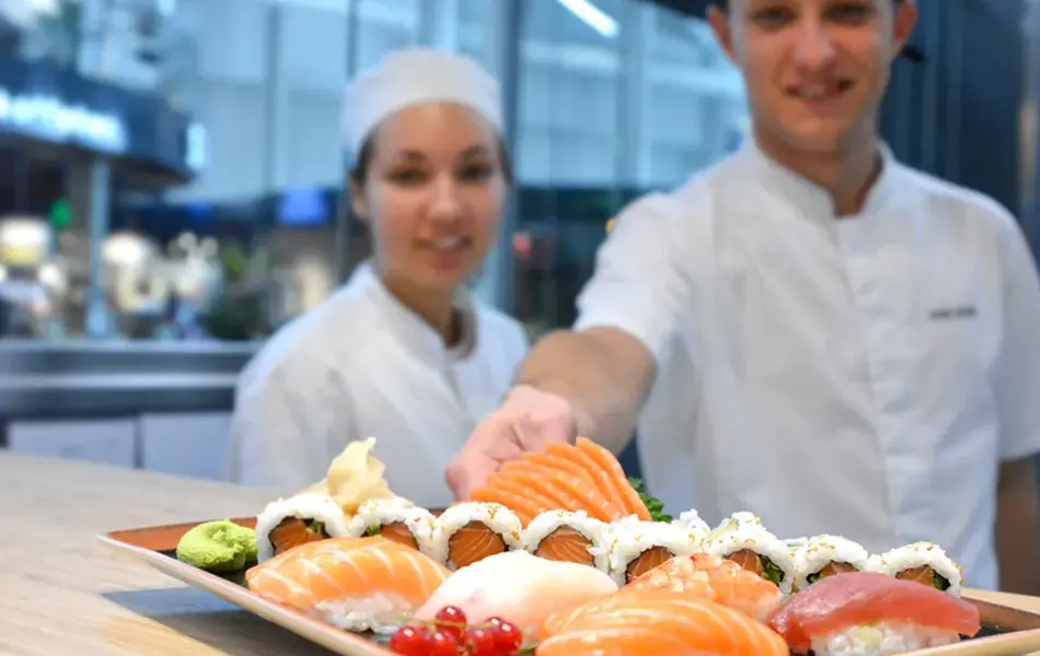 To sushi kokk som smiler og serverer sushi på benken