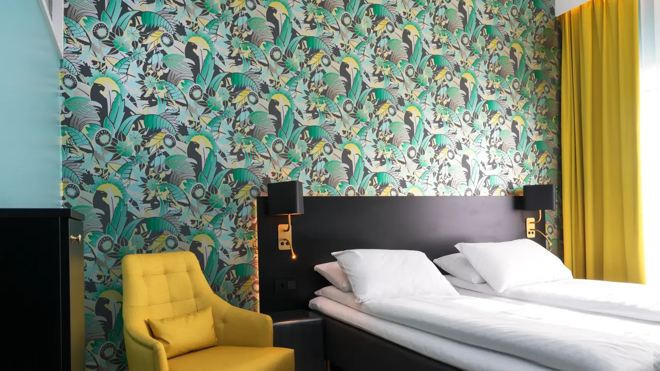 Bilde av standard dobbeltrom med dobbeltseng, gul lenestol, skap med strykebrett, mønstrete tapet i grønnt, gult , grått og sort, parkettgulv og gulv-til-tak-vindu med gule gardiner.